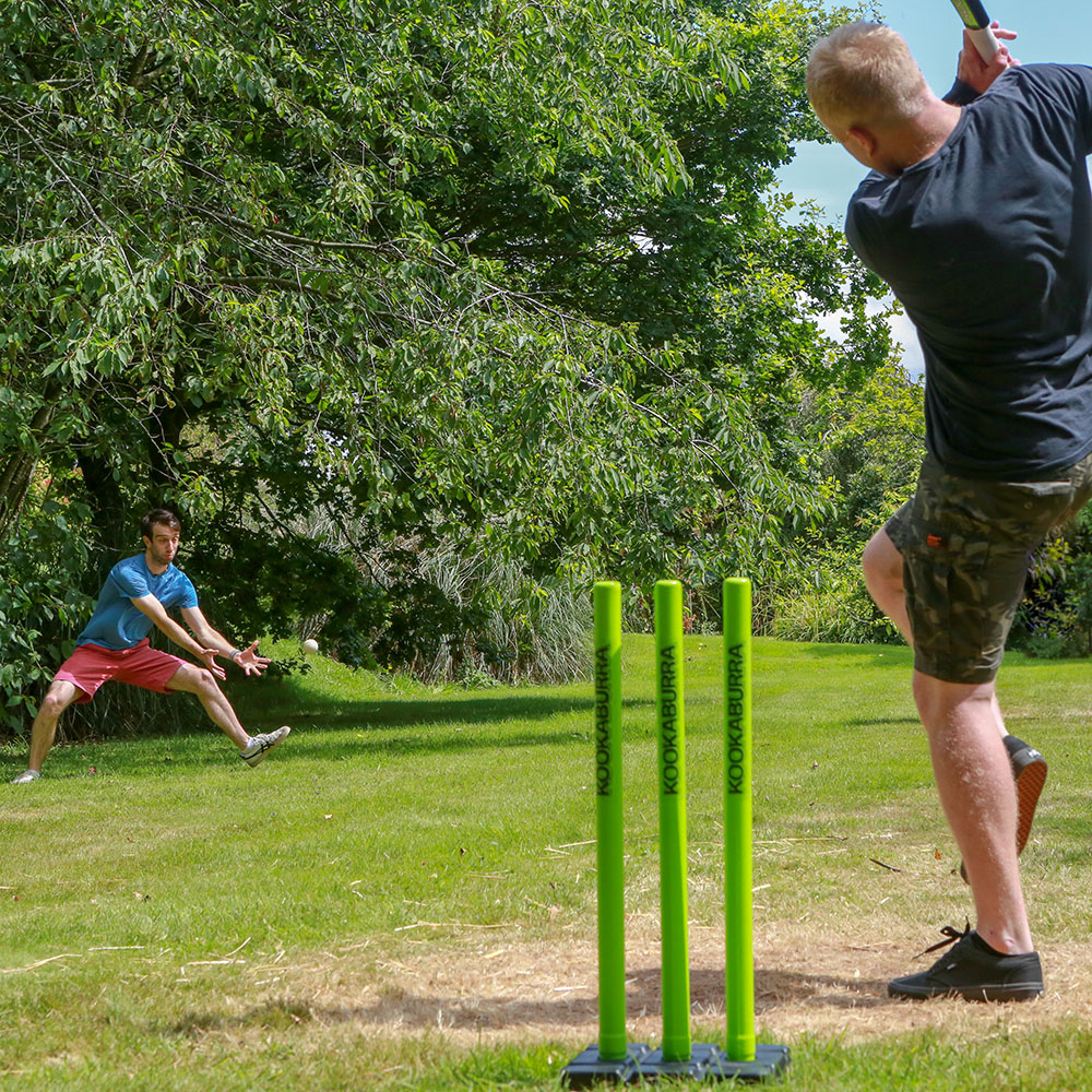 One man hitting a cricket ball across a garden and another about to catch it.